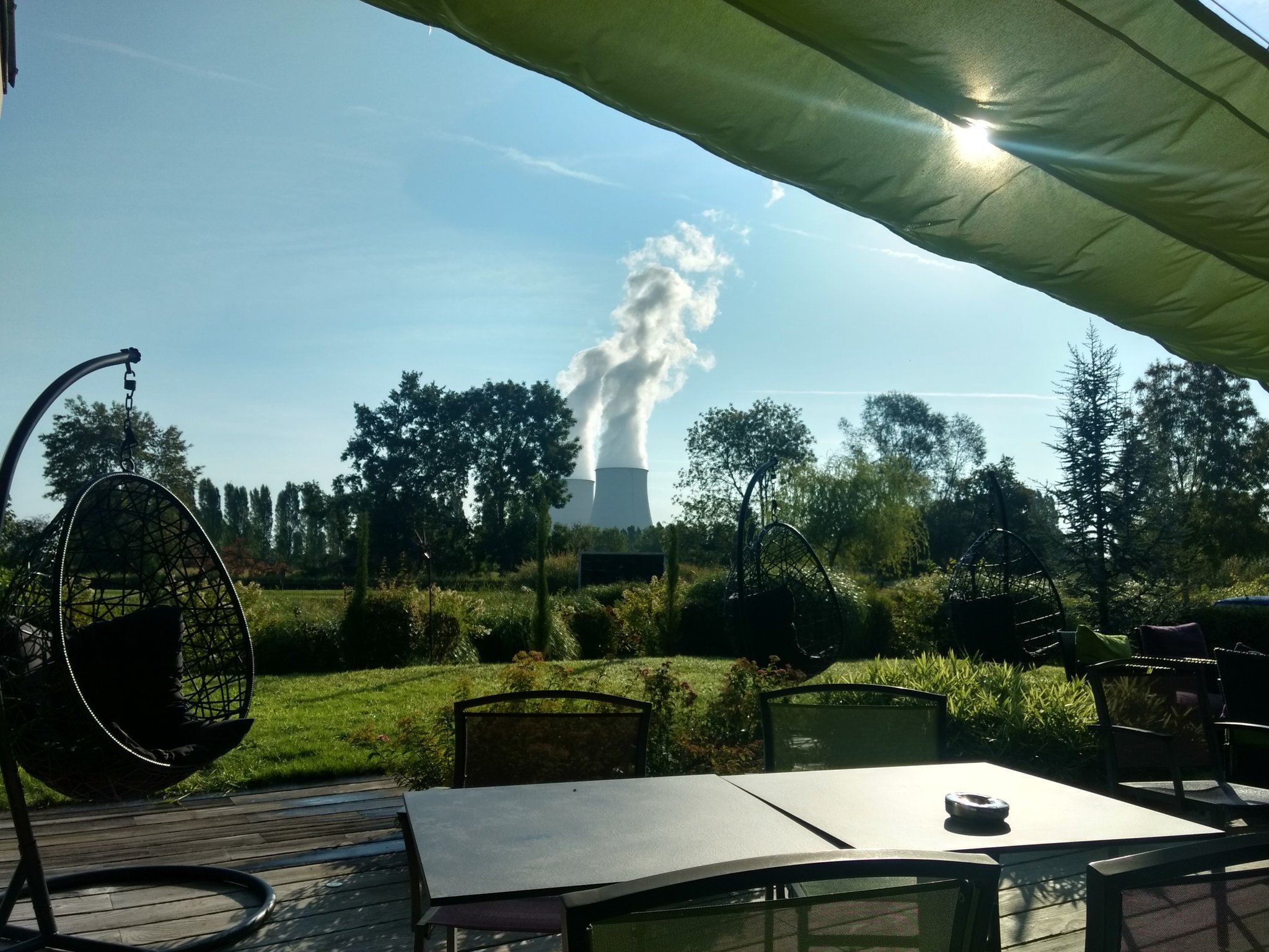 Photo d'une terrasse de restaurant ensoleillée, avec en arrière-plan un fauteuil en osier suspendu et une centrale nucléaire.