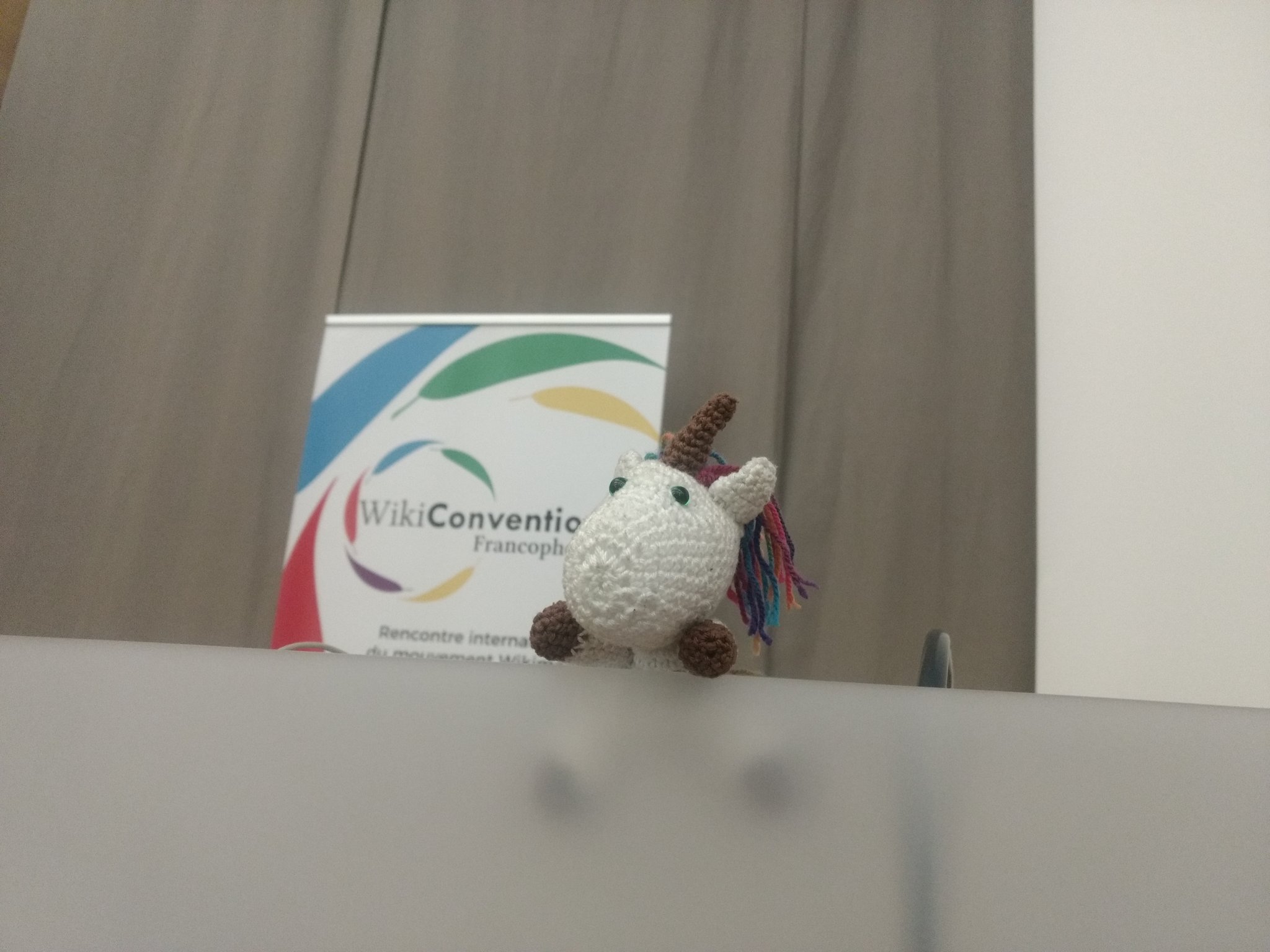 Peluche de licorne blanche au crochet sur un podium de salle de conférence. Derrière elle un dérouleur affiche « Wikiconvention francophone ».