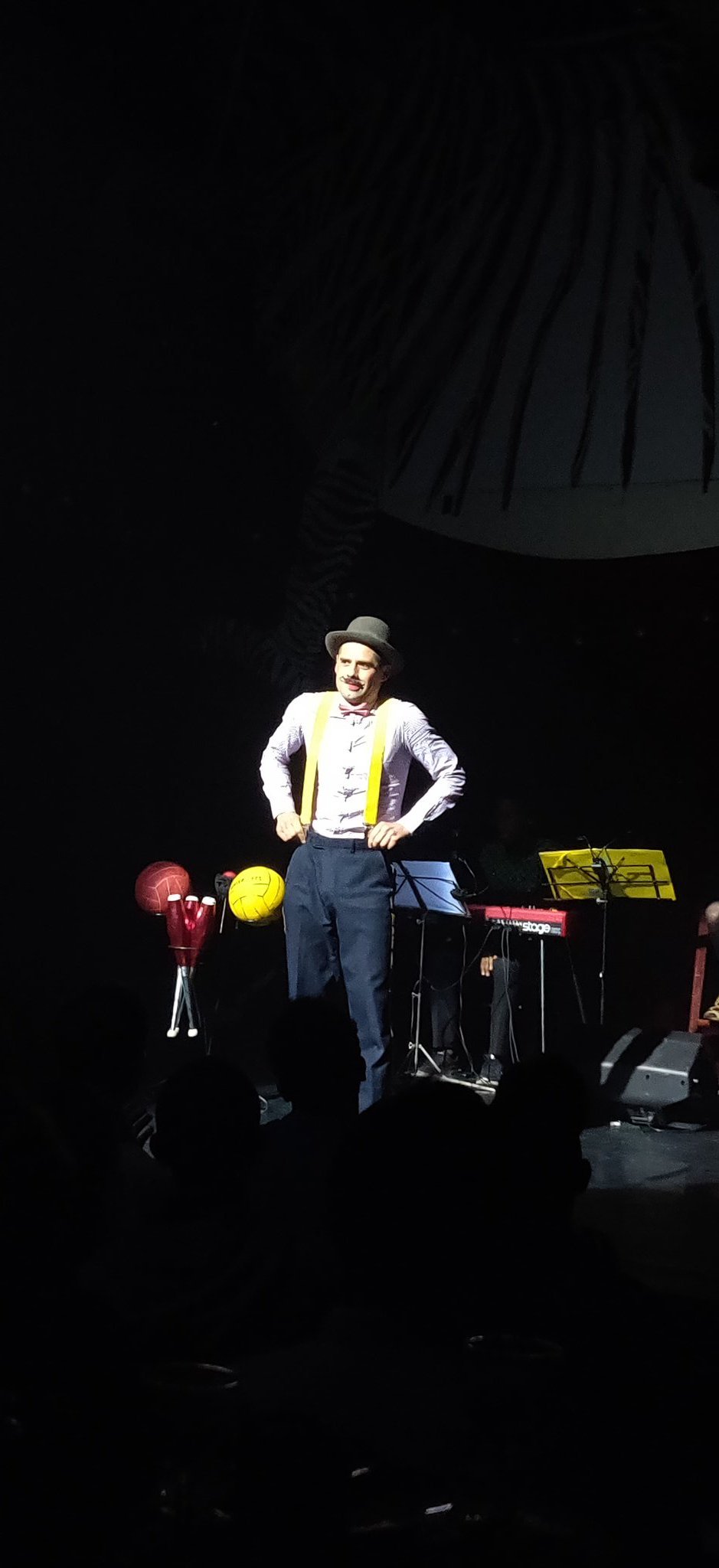 Un homme sur scène avec un chapeau gris et des bretelles jaunes. Un présentoir à côté de lui est chargé d’accessoires de jonglerie.