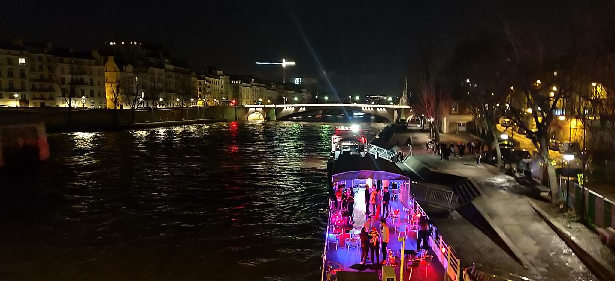 Photo de nuit prise sur un quai de la Seine. Au premier plan, une péniche pleine de lumières colorées.