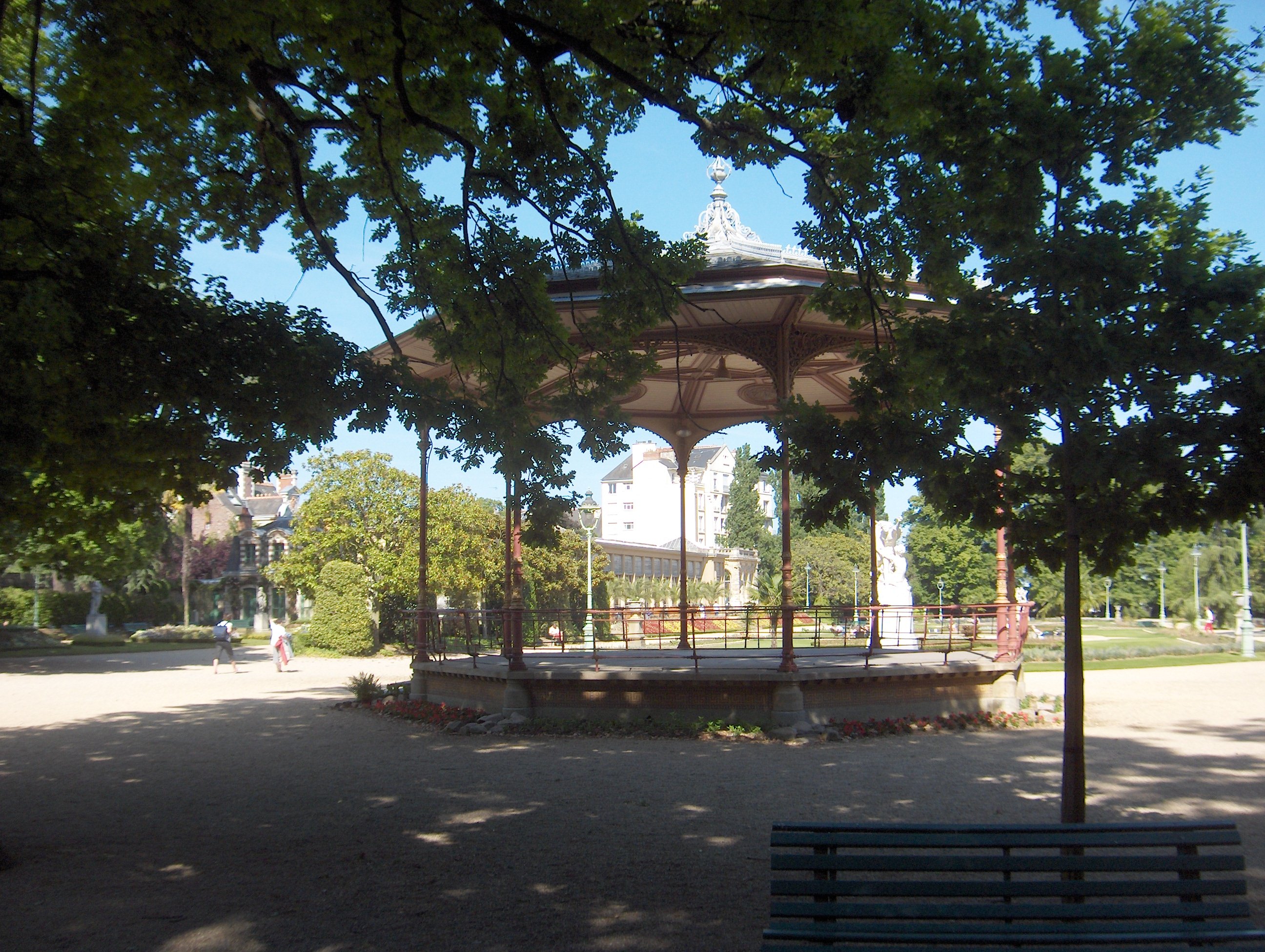 Un kiosque à musique en métal au milieu d'une étendue ouverte, avec deux arbres au premier plan.