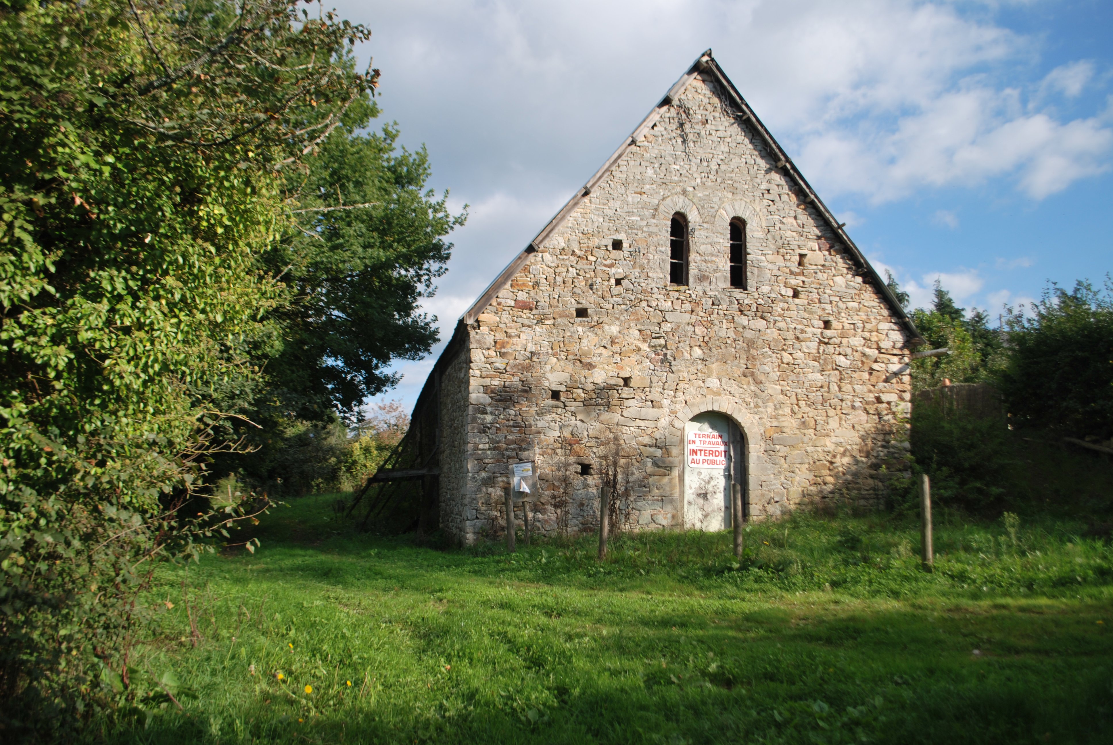 Façade d'une chapelle médiévale au milieu des bois. Une pancarte sur la porte dit « Terrain en travaux. Interdit au public »