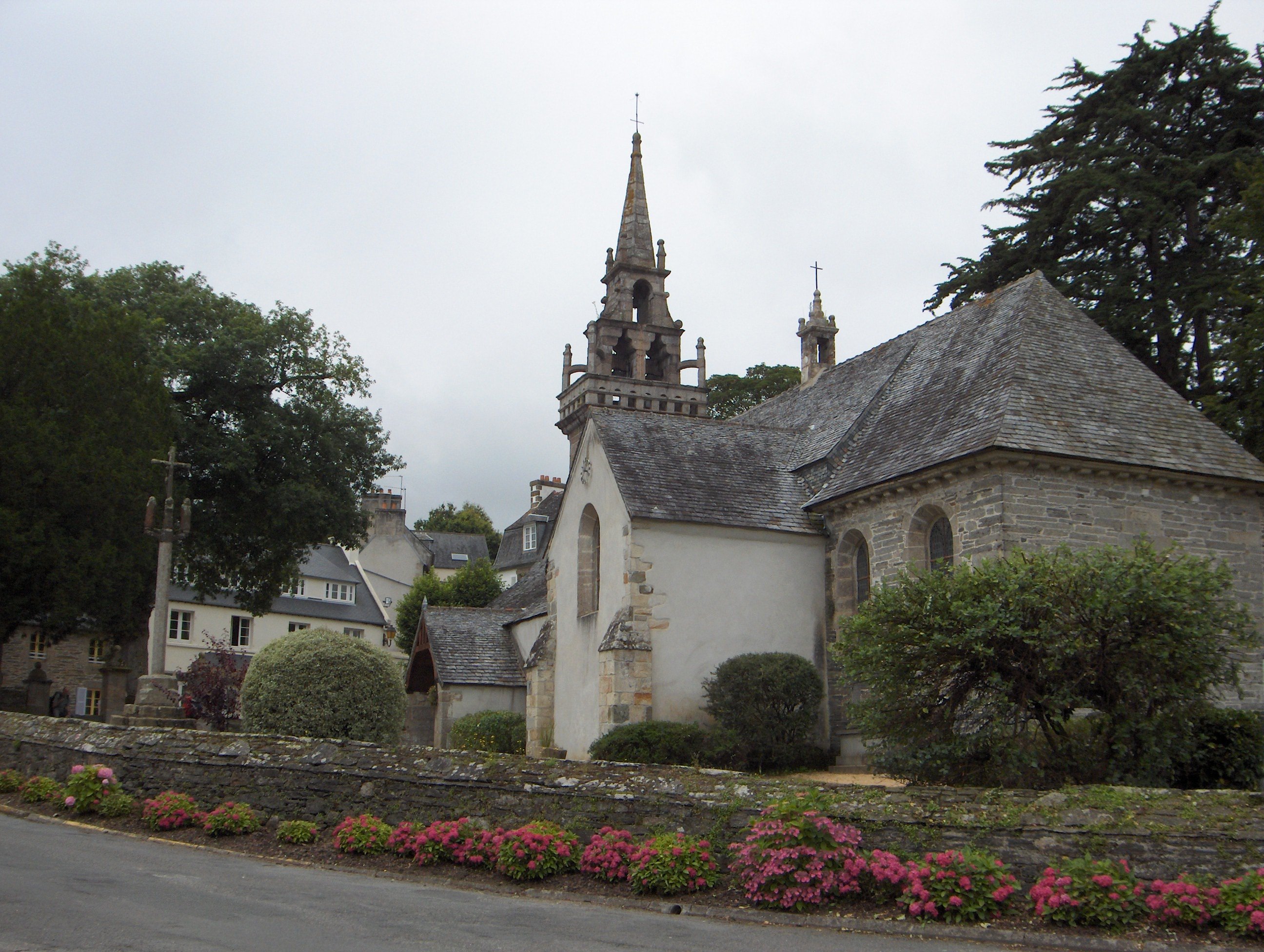 Une petite église gothique avec un clocher ouvrage, un calvaire sur le côté et un muret la séparant de la route.