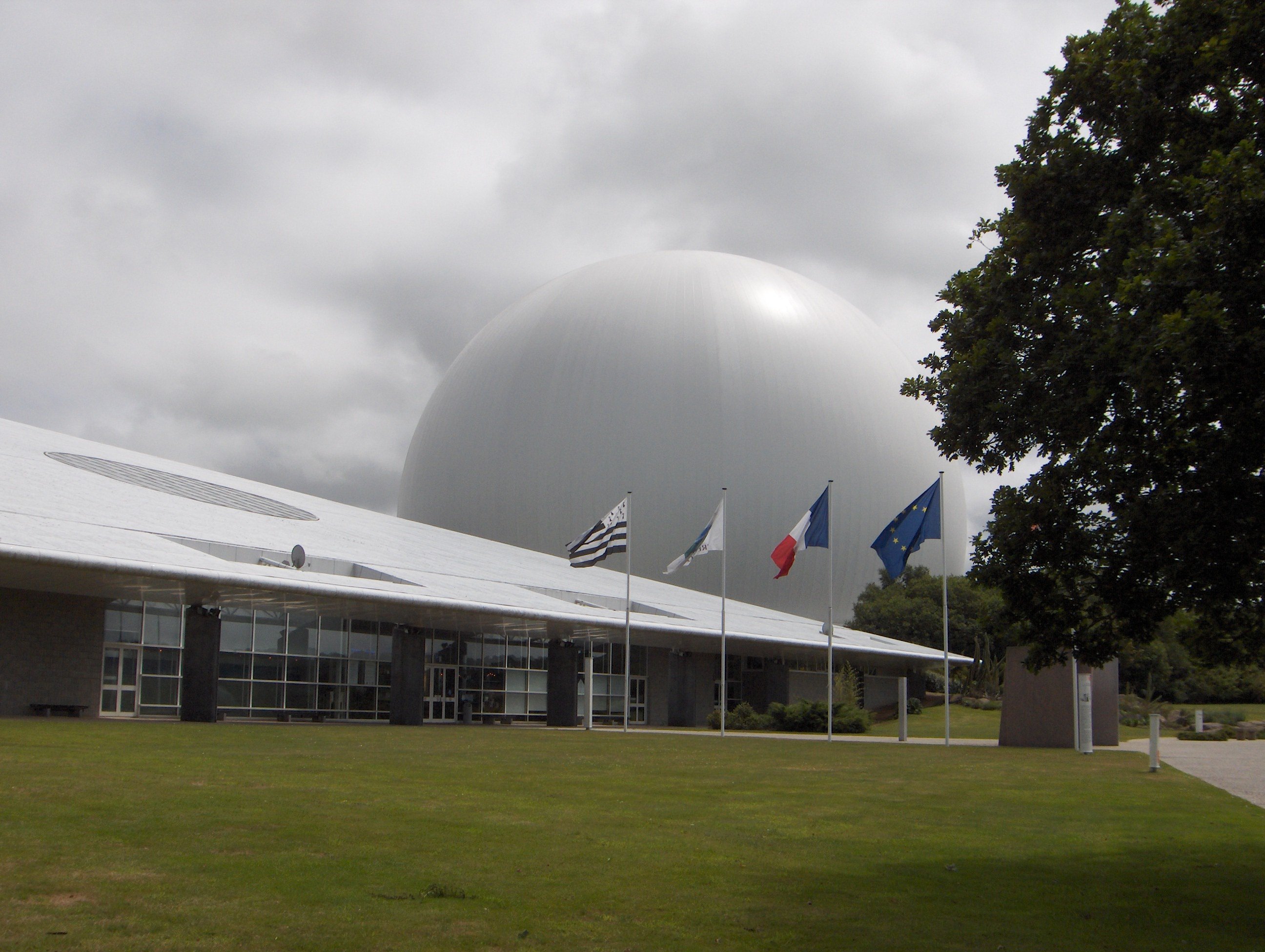 Une énorme sphère blanche dépasse devant un bâtiment triangulaire, avec quatre drapeaux devant, dont les drapeaux breton, français et européen ainsi qu'un quatrième non-identifiable ?