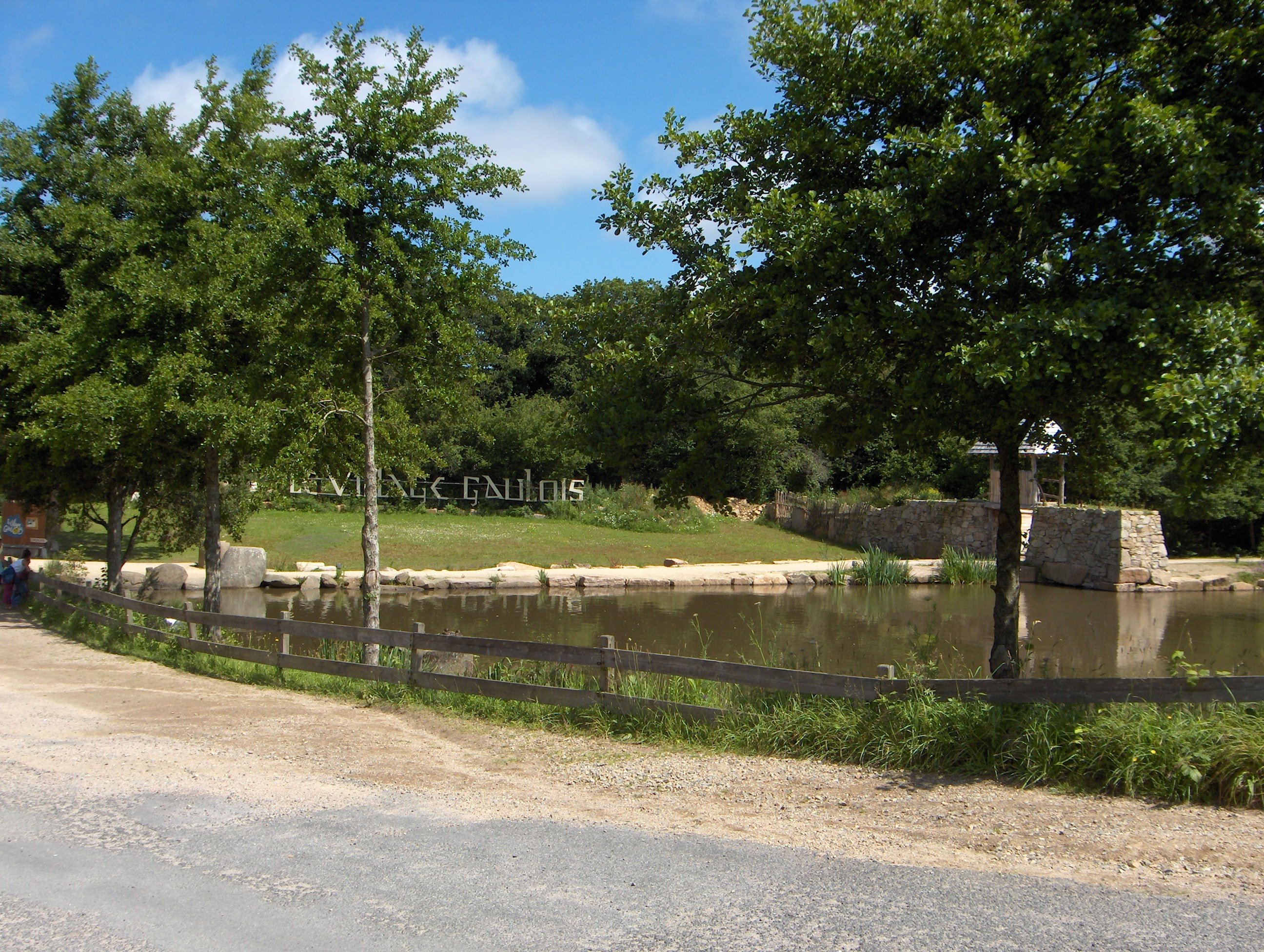 Une allée bordée d'arbres devant un étang. De l'autre côté de celui-ci, des lettres géantes disposées verticalement forment les mots "Le Village gaulois"