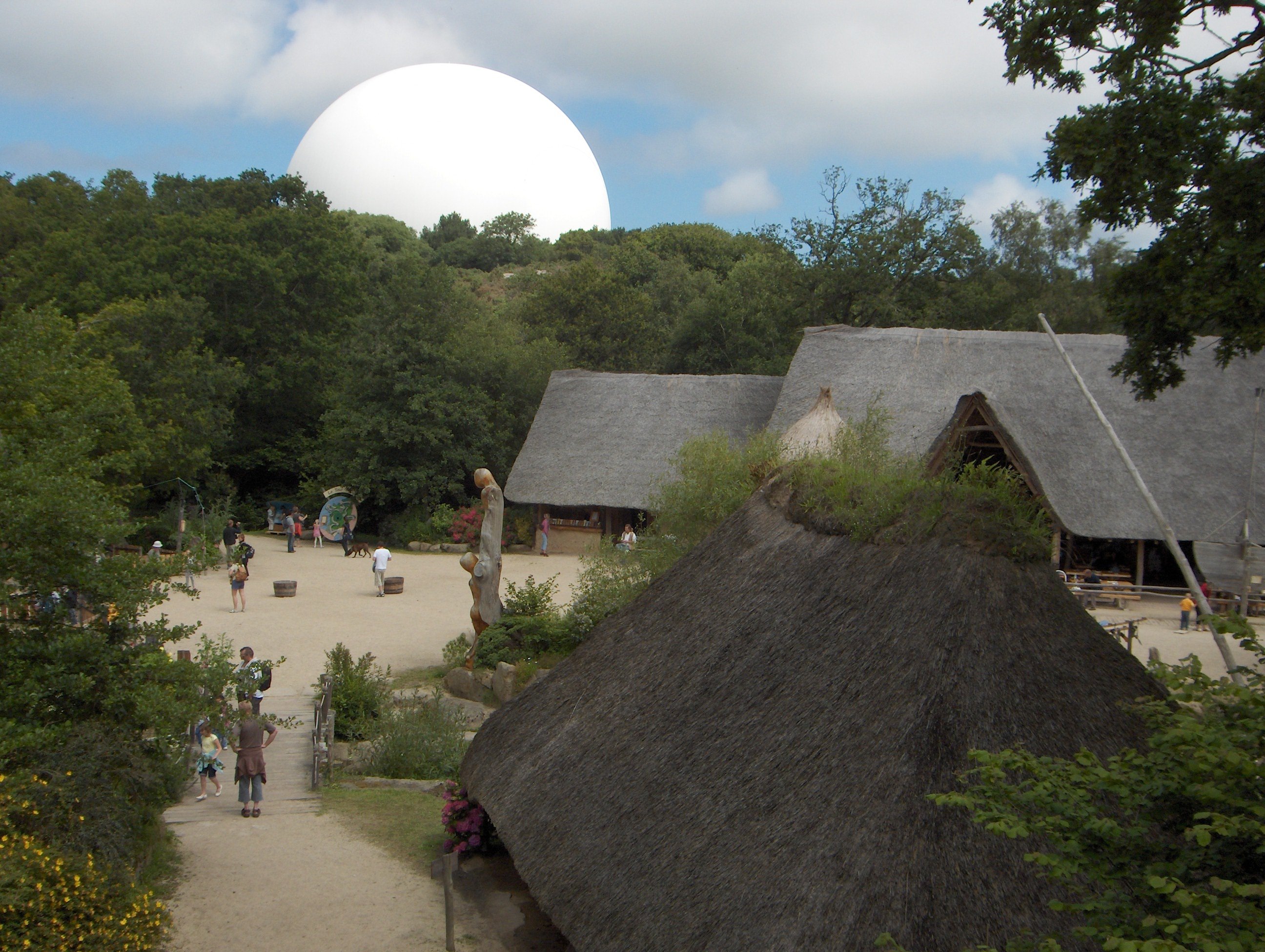 Des bâtiments en bois avec un toit en chaume au milieu d'une clairière. avec une statue en bois représentant une silhouette. Derrière les arbres, la sphère blanche du Radôme dépasse de façon incongrue.
