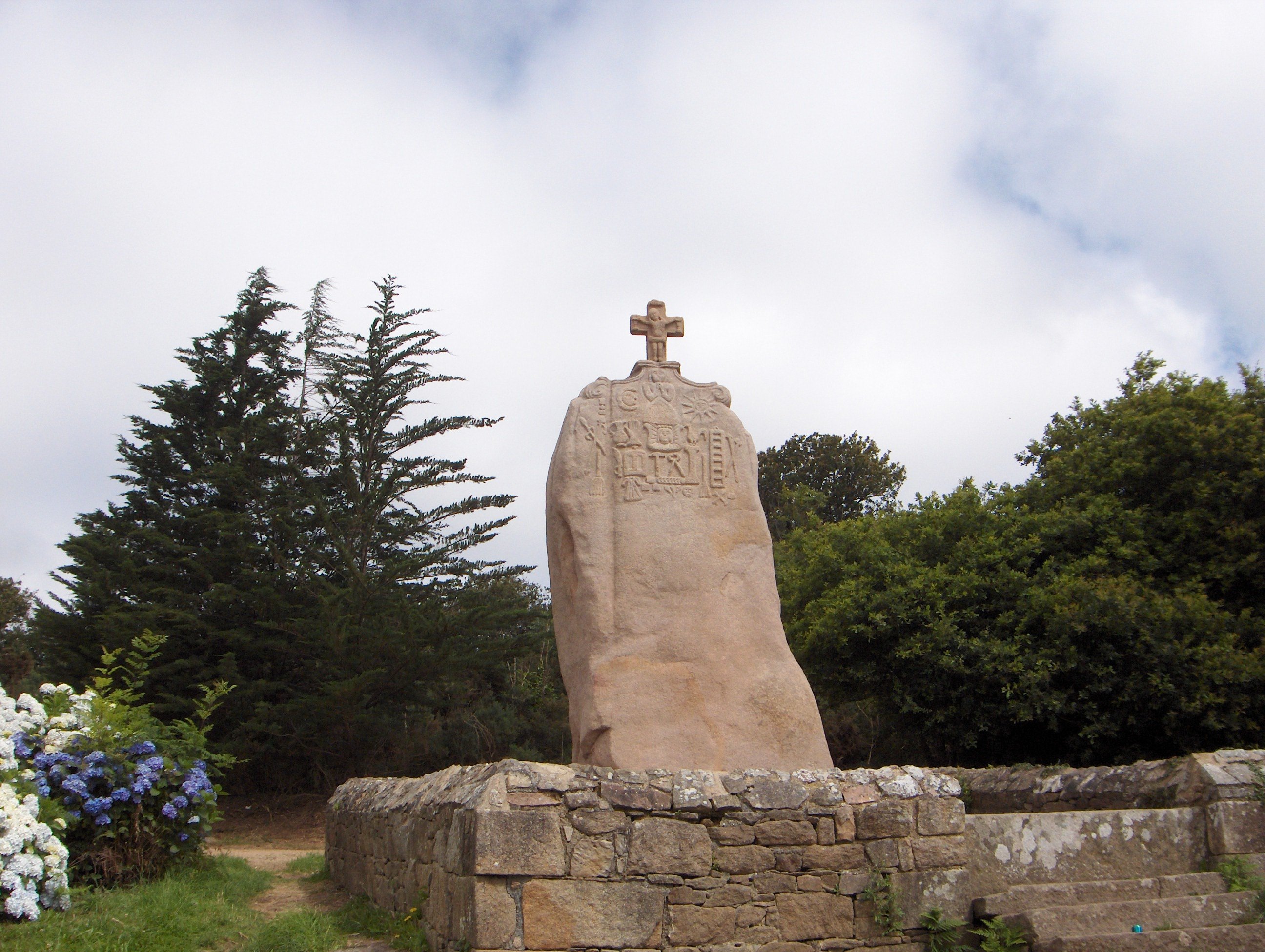 Un menhir entouré d'un muret de pierre. Un crucifix a été ajouté au sommet, et la surface est partiellement couverte de gravures.