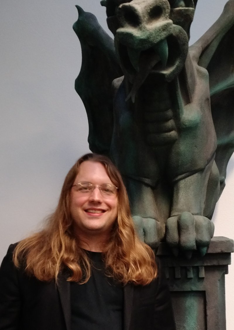 Portrait d'un homme avec de longs cheveux blonds-roux, posé devant une statue de dragon. Il porte des lunettes et sourit.