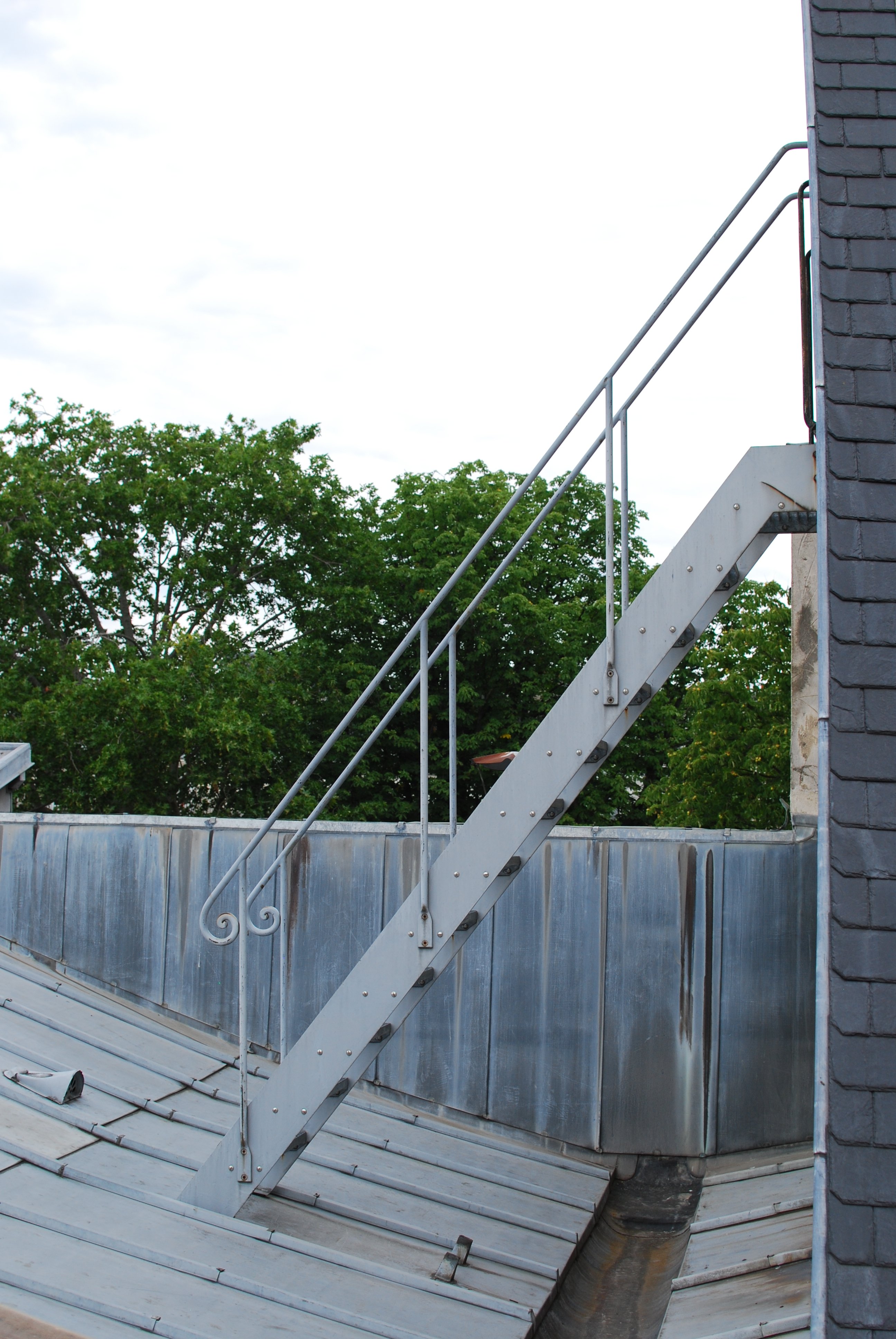 Escalier en métal vu de profil, émergeant du côté d'un bâtiment couvert d'ardoises, et descendant vers un toit en tôle presque plat.