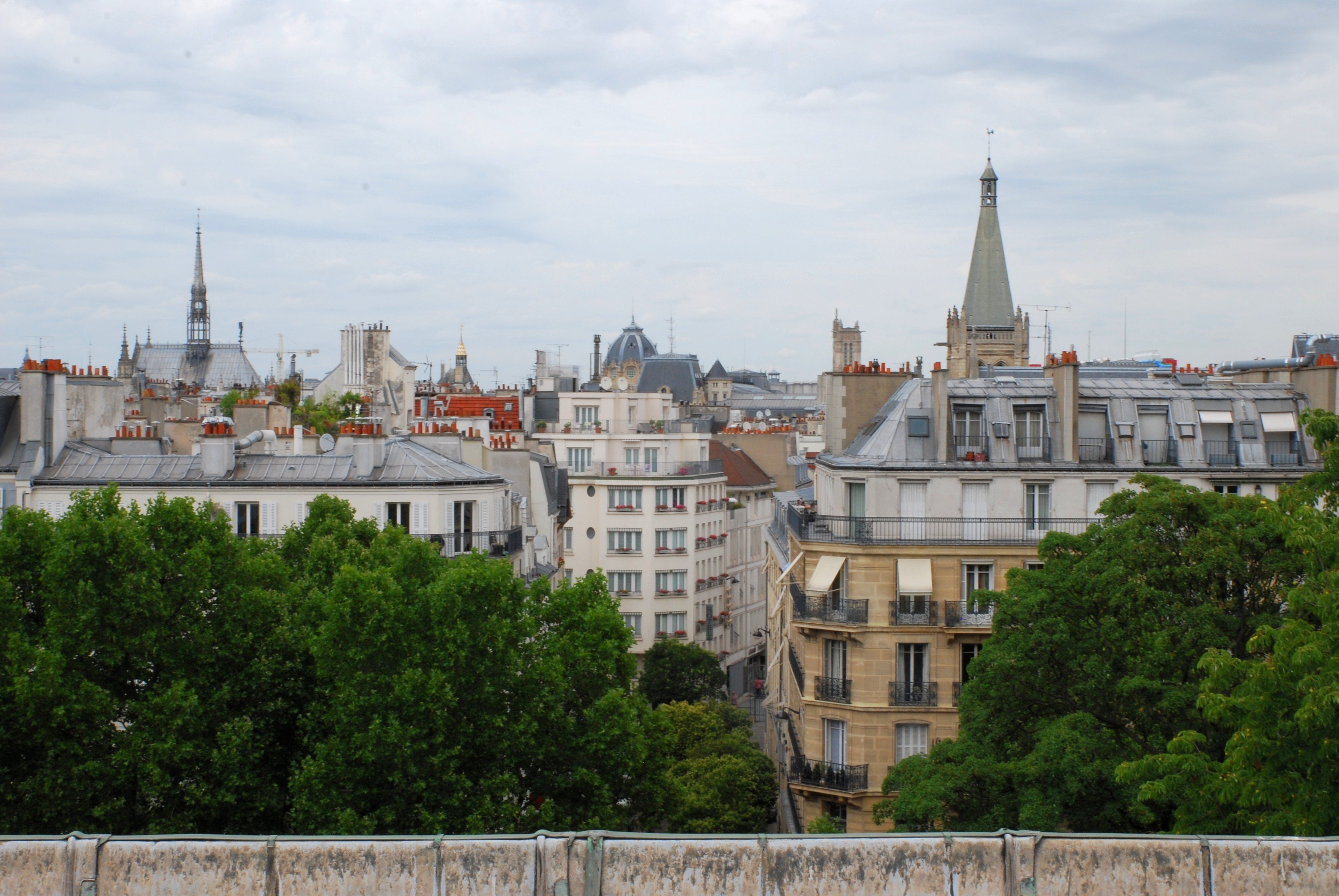 Des immeubles parisiens au-delà d'un rideau d'arbres, avec quelques édifices qui dépassent : la Sainte-Chapelle sur la gauche, l'église Saint-Séverin sur la droite et le dôme du tribunal de commerce de Paris entre les deux.