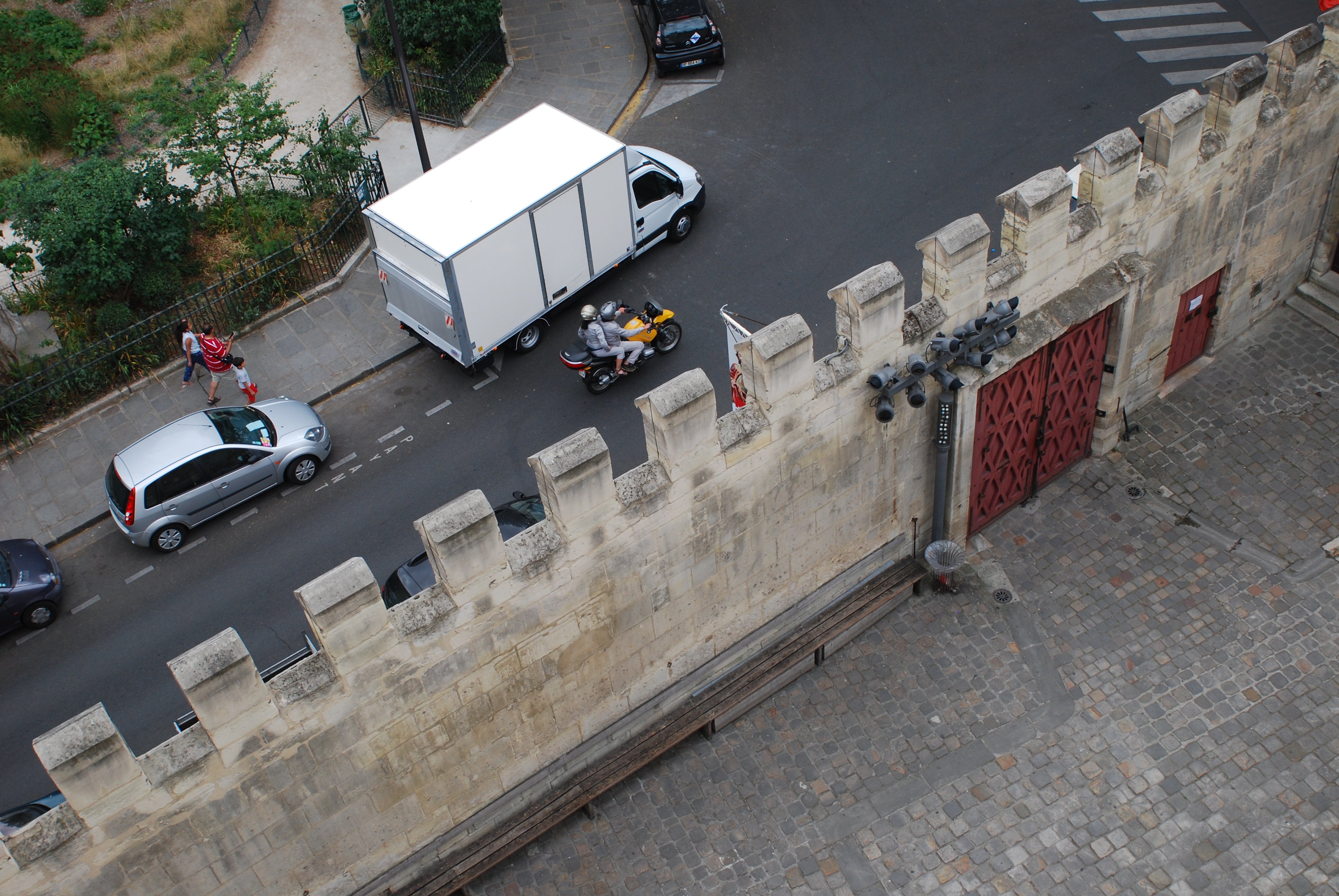 Photo délimitée en deux par le mur de la cour de l’hôtel de Cluny. En haut à gauche, la rue, avec des véhicules stationnés, une moto jaune sur la chaussée et des passants sur le trottoir. En bas à droite, le mur lui-même, crénelé et percé de deux portes anciennes couleur lie-de-vin, et la cour pavée.