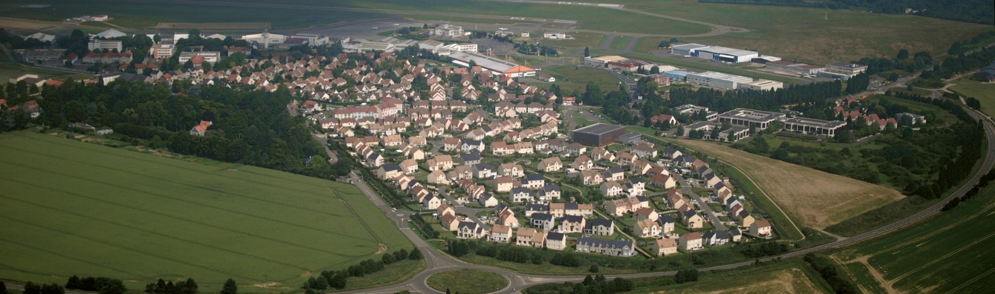 Une petite ville formant un ensemble assez dense de maisons individuelles entourées de champs. On voit l’aéroport au fond.