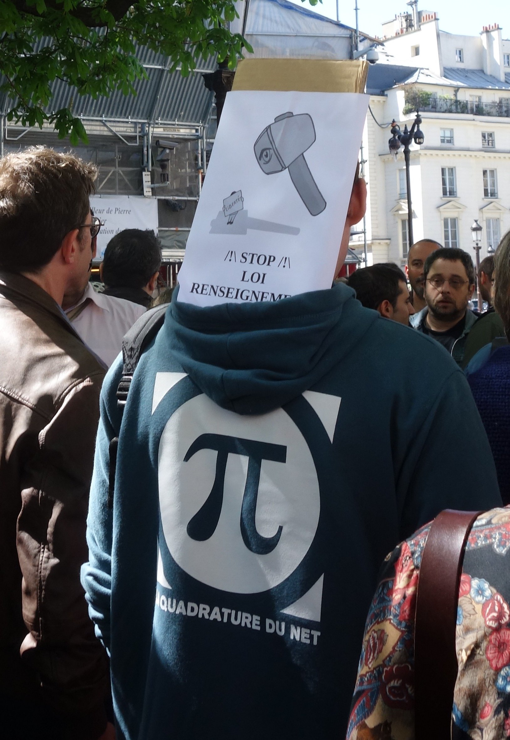 Un homme vu de dos, portant un hoodie bleu avec le logo de la Quadrature du Net duquel dépasse une pancarte dépeignant un marteau doté d'un œil prêt à écraser un petit bonhomme caché derrière une pancarte "liberté". En dessous figure la mention "/!\ STOP /!\  LOI RENSEIGNEMENT"