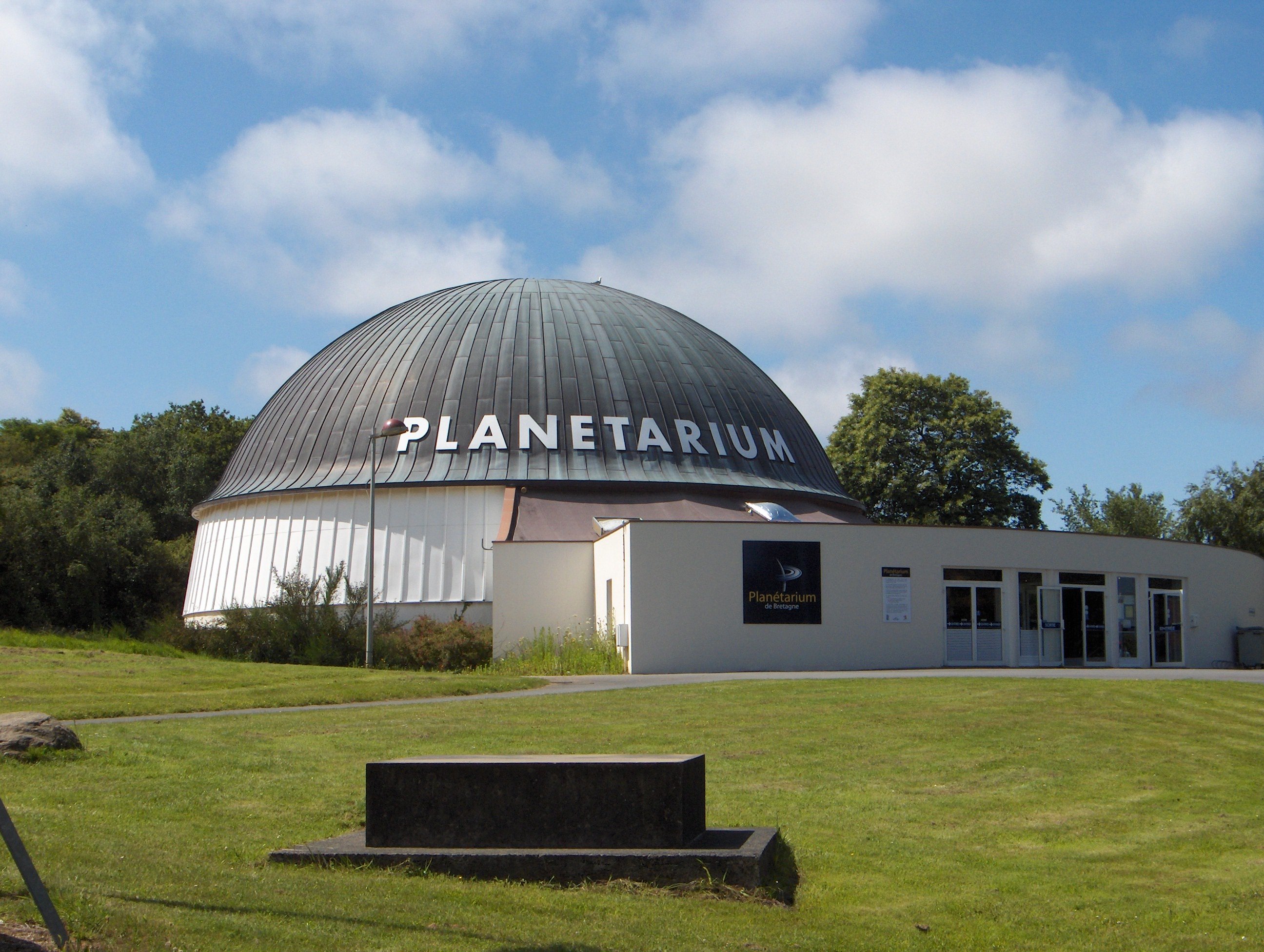 Un bâtiment en forme de cylindre blanc surmonté d’un dôme vert-de-gris, avec le mot "PLANETARIUM" écrit en blanc.