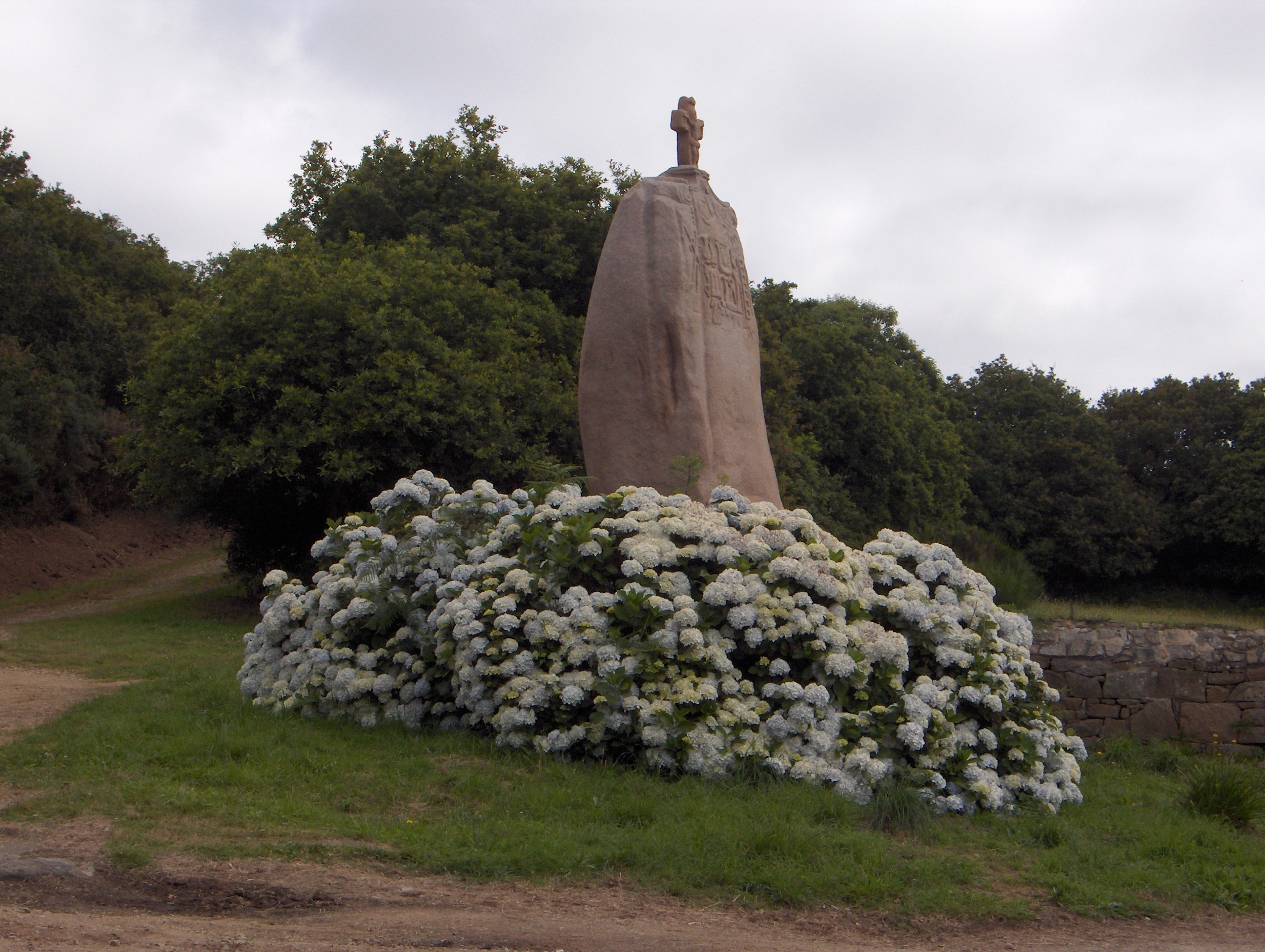 Un menhir entouré d'un muret de pierre. Un crucifix a été ajouté au sommet, et la surface est partiellement couverte de gravures. Un massif de fleurs blanches se trouve au premier plan.