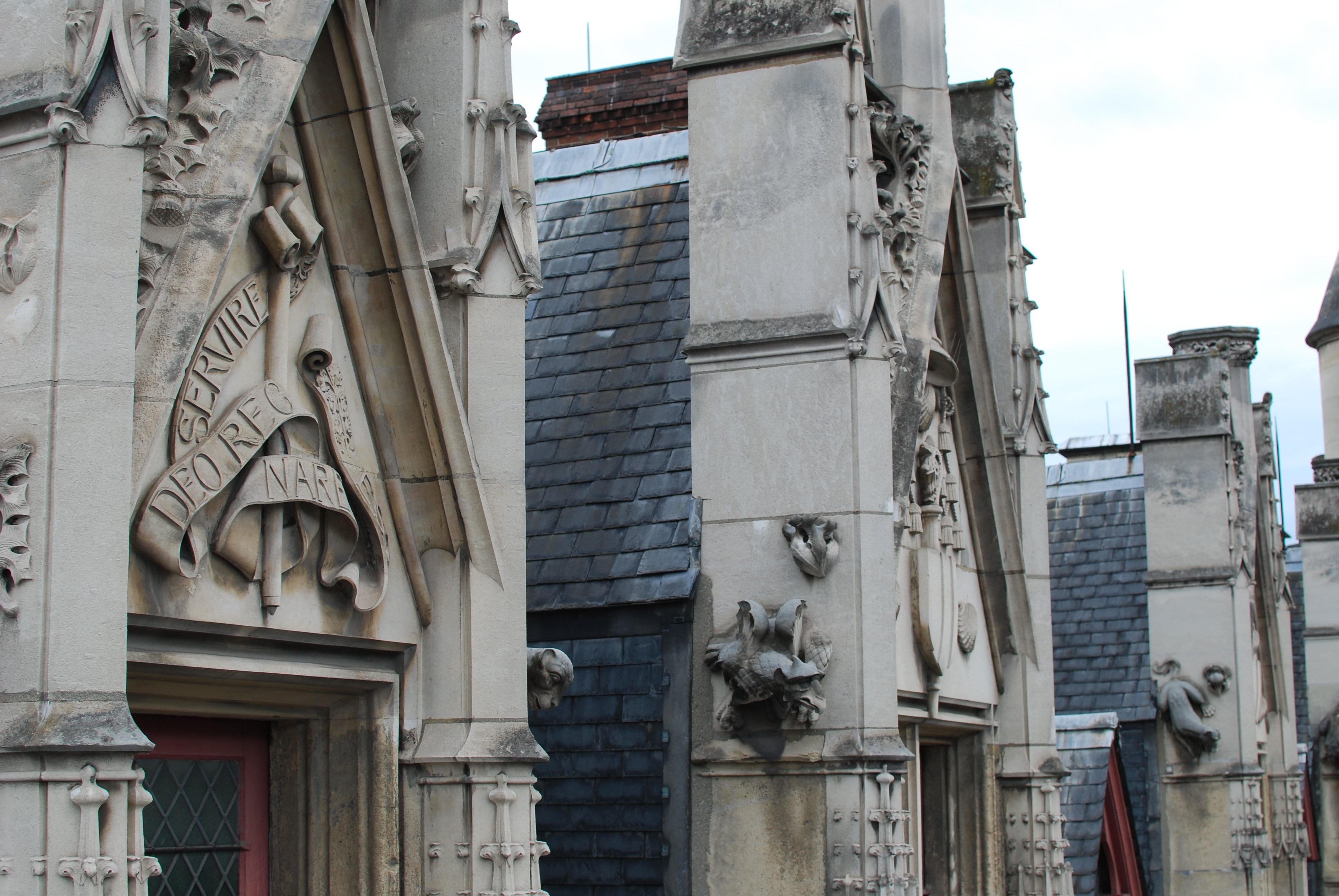 Détail des encadrements de fenêtre au niveau des toits. Elles sont surmontées d'un fronton triangulaire en pierre où figurent des écus ou des devises en latin. Les bords supérieurs du fronton portent des motifs de feuille de houx, et des grotesques émergent des montants verticaux sur les côtés.