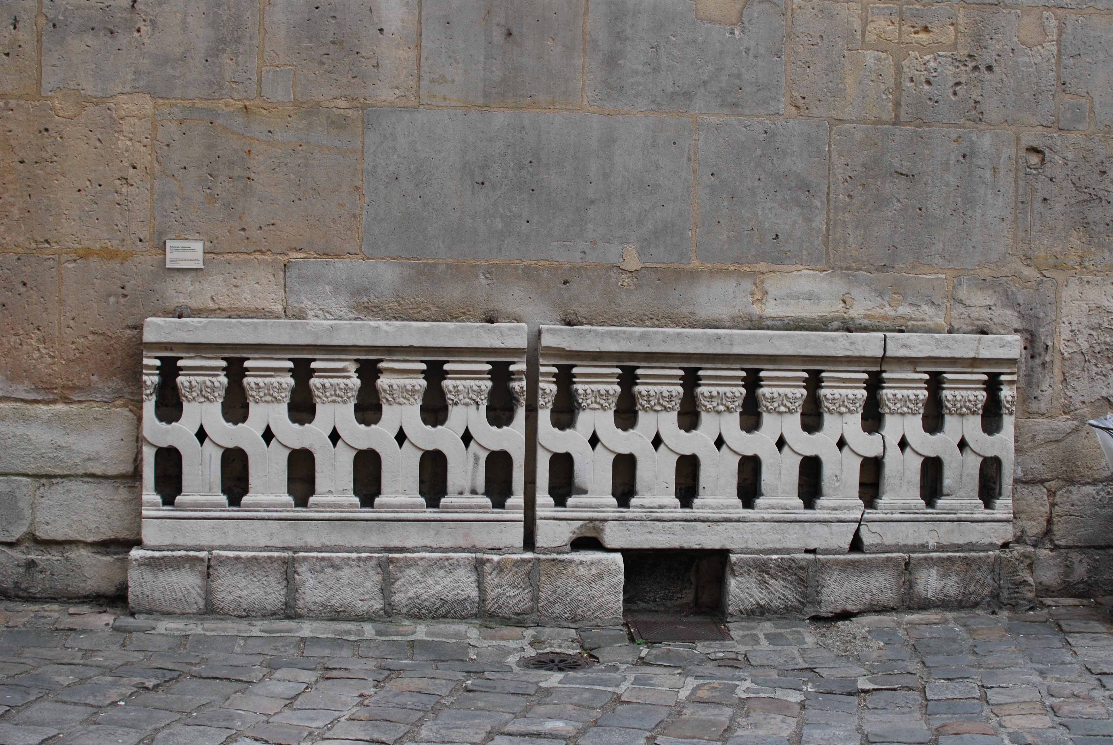 Une balustrade en pierre posée devant un mur. Un cartel juste au-dessus contient la description qui accompagne cette image.